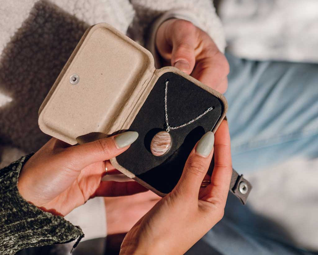 An open kraft jewelry gift box with a custom stone necklace inside, held by a couple