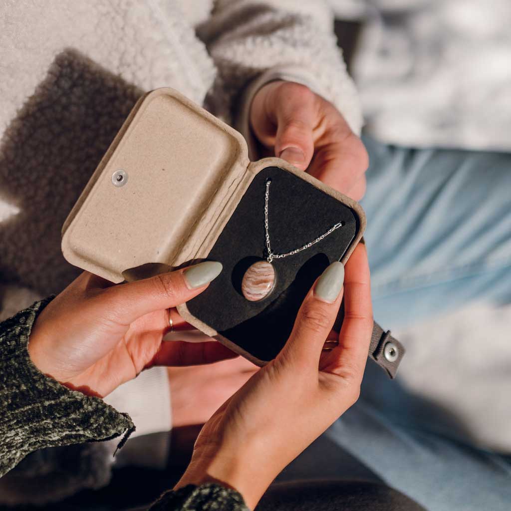 An open kraft jewelry gift box with a custom stone necklace inside, held by a couple