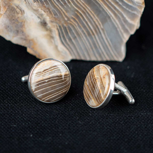 Striped chert cufflinks on a dark textured surface with the stone they were made from in the background