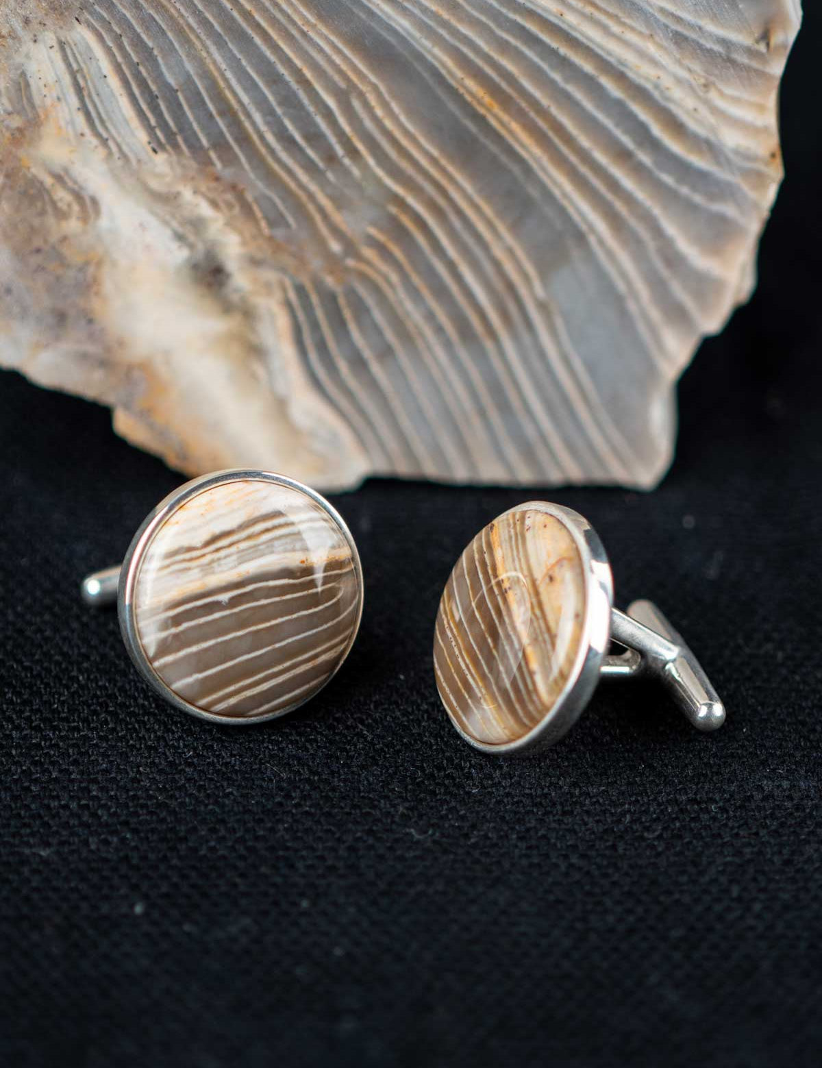 Striped chert cufflinks on a dark textured surface with the stone they were made from in the background
