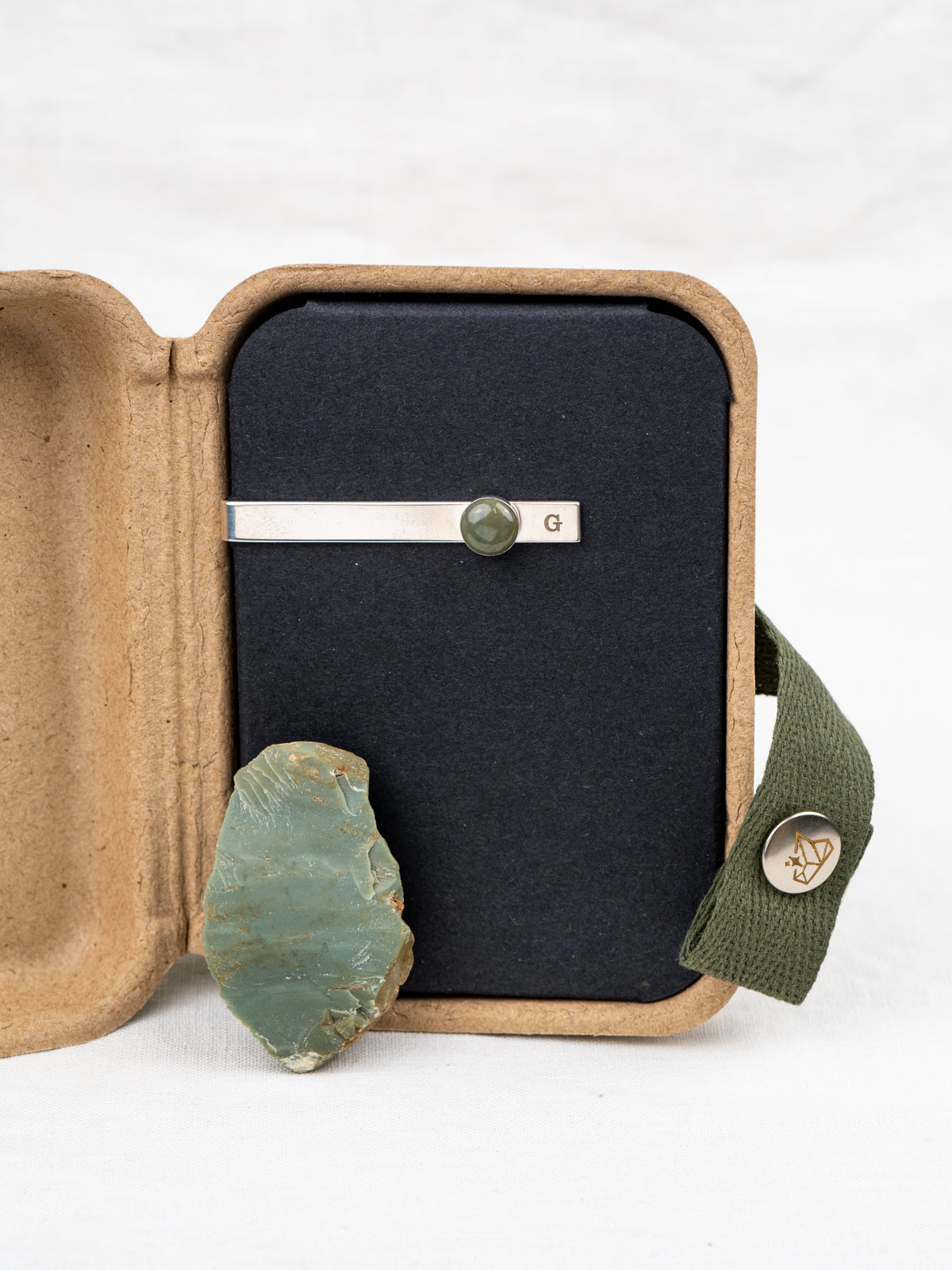 Sterling silver tie clip with a mounted round green stone, displayed in a kraft gift box with the original rock it was made from
