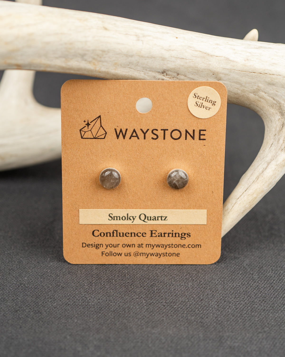 Waystone smoky quartz confluence stud earrings on a kraft paper card with antlers in the background