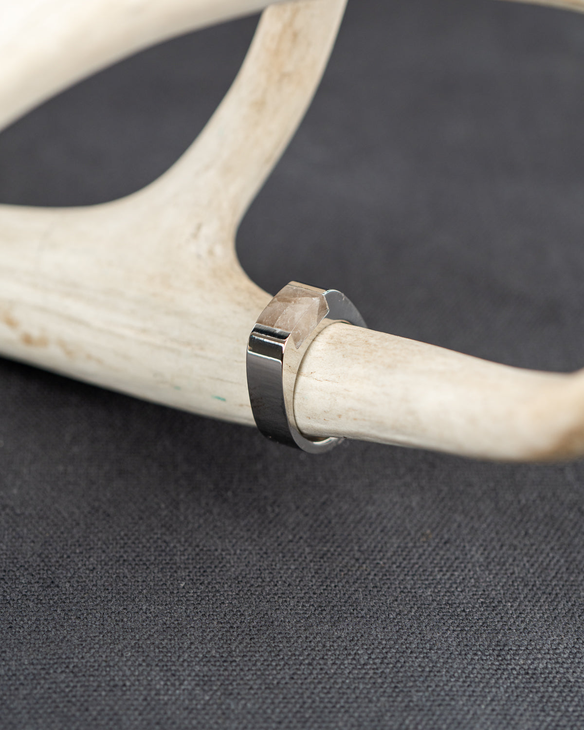 A chunky stainless steel ring set with a smoky quartz stone rests on an antler against a gray background