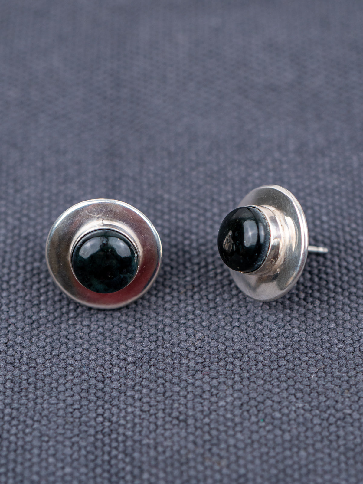 Pair of sterling silver stud earrings featuring round black jade stones, photographed on a textured gray fabric background, shown from front and side angles.