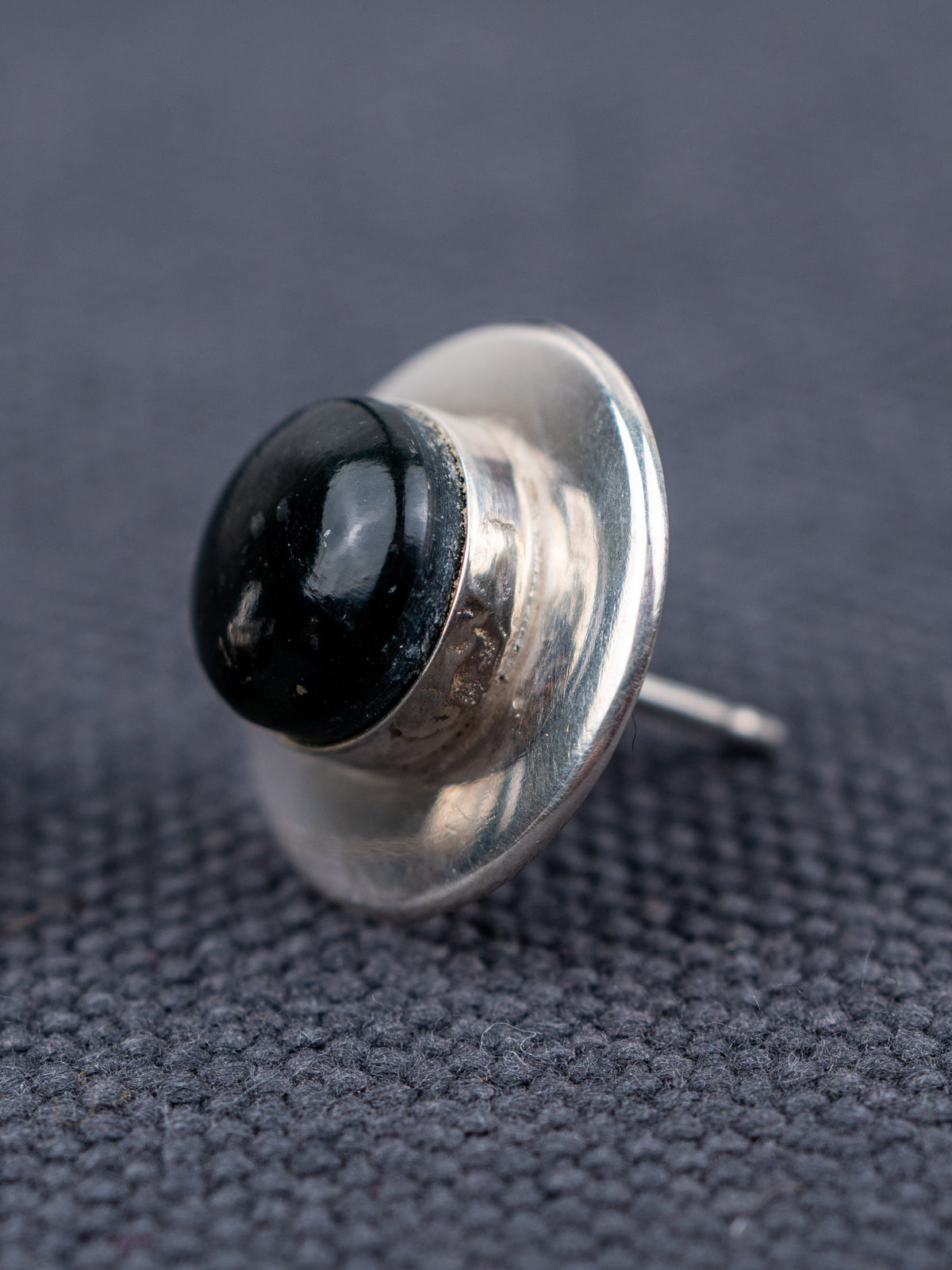 Sterling silver stud earring with a domed black jade stone, shown in close-up from a side angle on a textured gray fabric background.