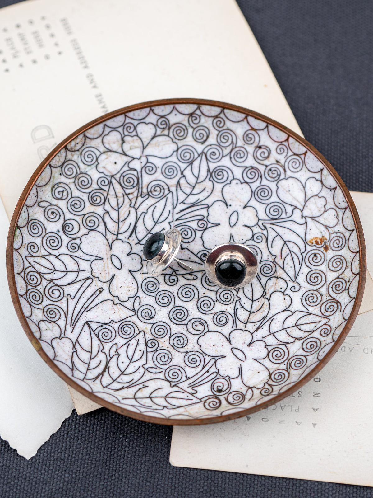 Pair of sterling silver black jade stud earrings displayed on a decorative ceramic dish with floral patterns, styled on layered paper over a dark fabric background.