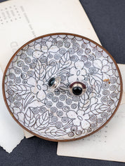 Pair of sterling silver black jade stud earrings displayed on a decorative ceramic dish with floral patterns, styled on layered paper over a dark fabric background.
