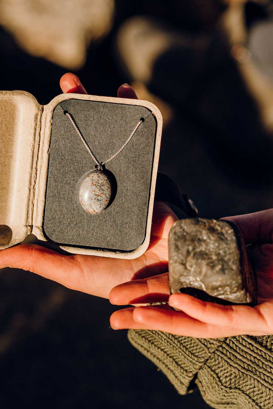 A woman holds an open kraft gift box displaying a natural stone pendant necklace in one hand, and the original stone the necklace was made from in the other hand