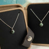 Two matching necklaces with a green stone pendant on a black display card inside a brown jewelry box.
