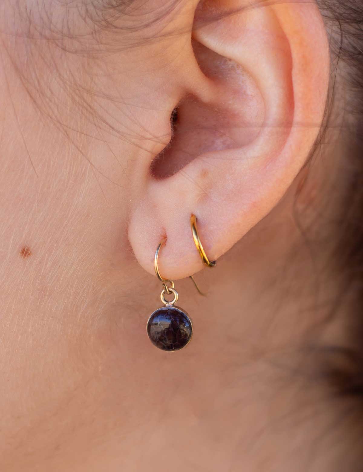 Close-up of an ear wearing a 14k gold fill dangling earring set with a natural dark stone. It&#39;s layered with a gold hoop earring.