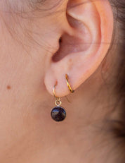Close-up of an ear wearing a 14k gold fill dangling earring set with a natural dark stone. It's layered with a gold hoop earring.