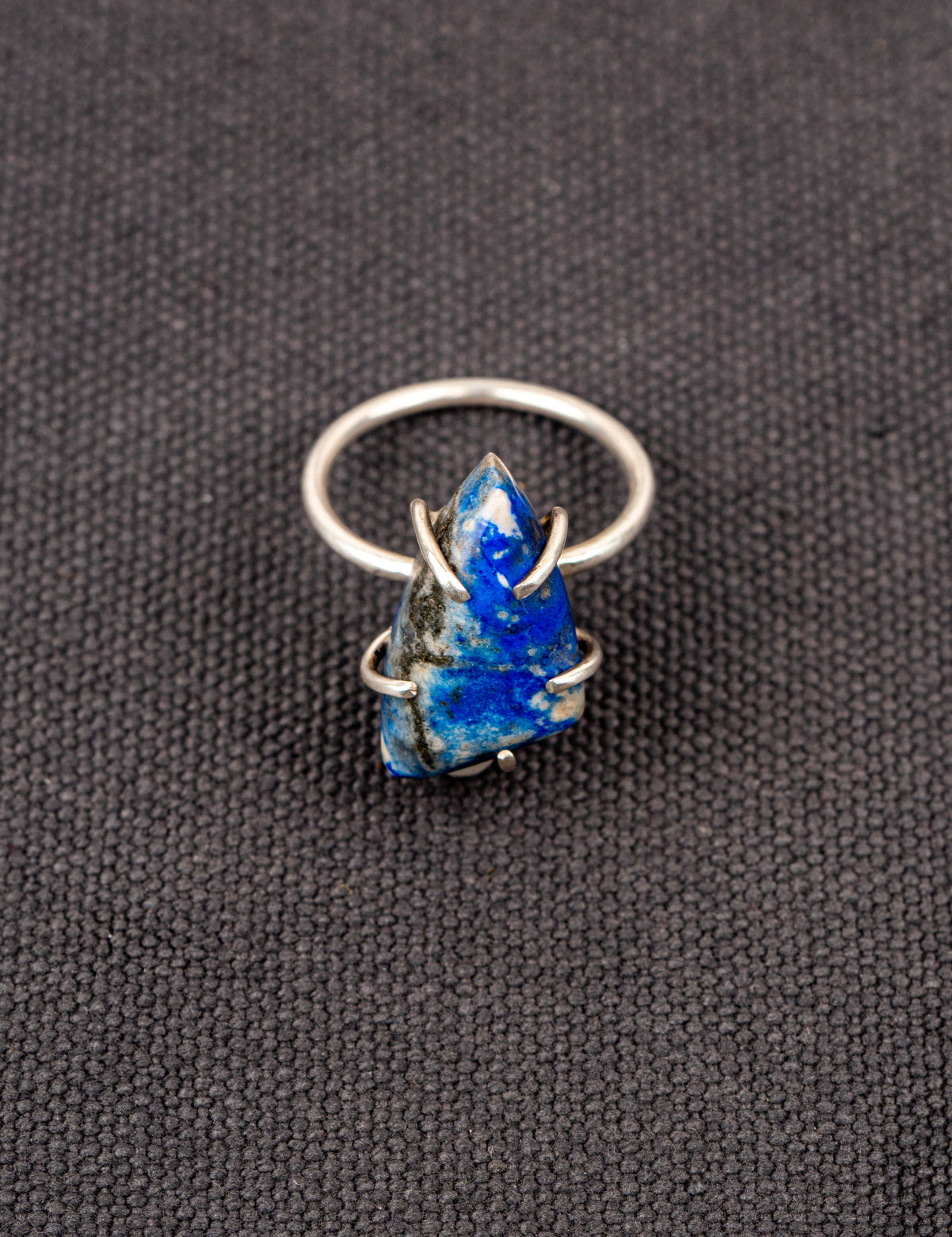 Silver ring with a deep blue lapis stone held securely by a prong setting, displayed on a dark fabric background.