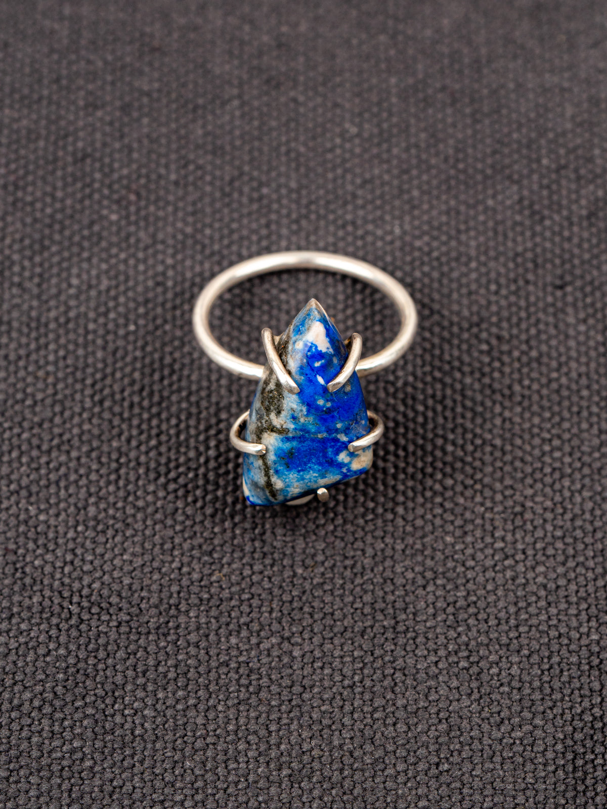 Silver ring with a deep blue lapis stone held securely by a prong setting, displayed on a dark fabric background.