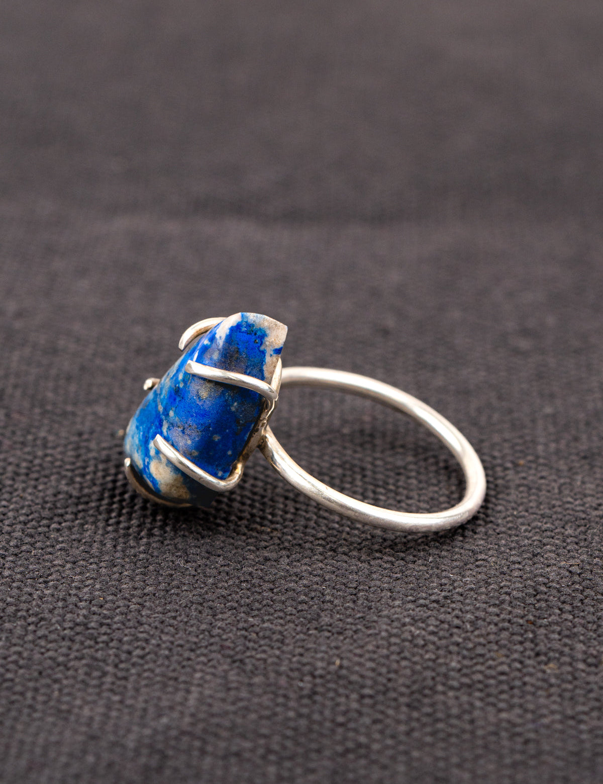 Side profile of a silver ring with a deep blue lapis stone held securely by a prong setting, displayed on a dark fabric background.