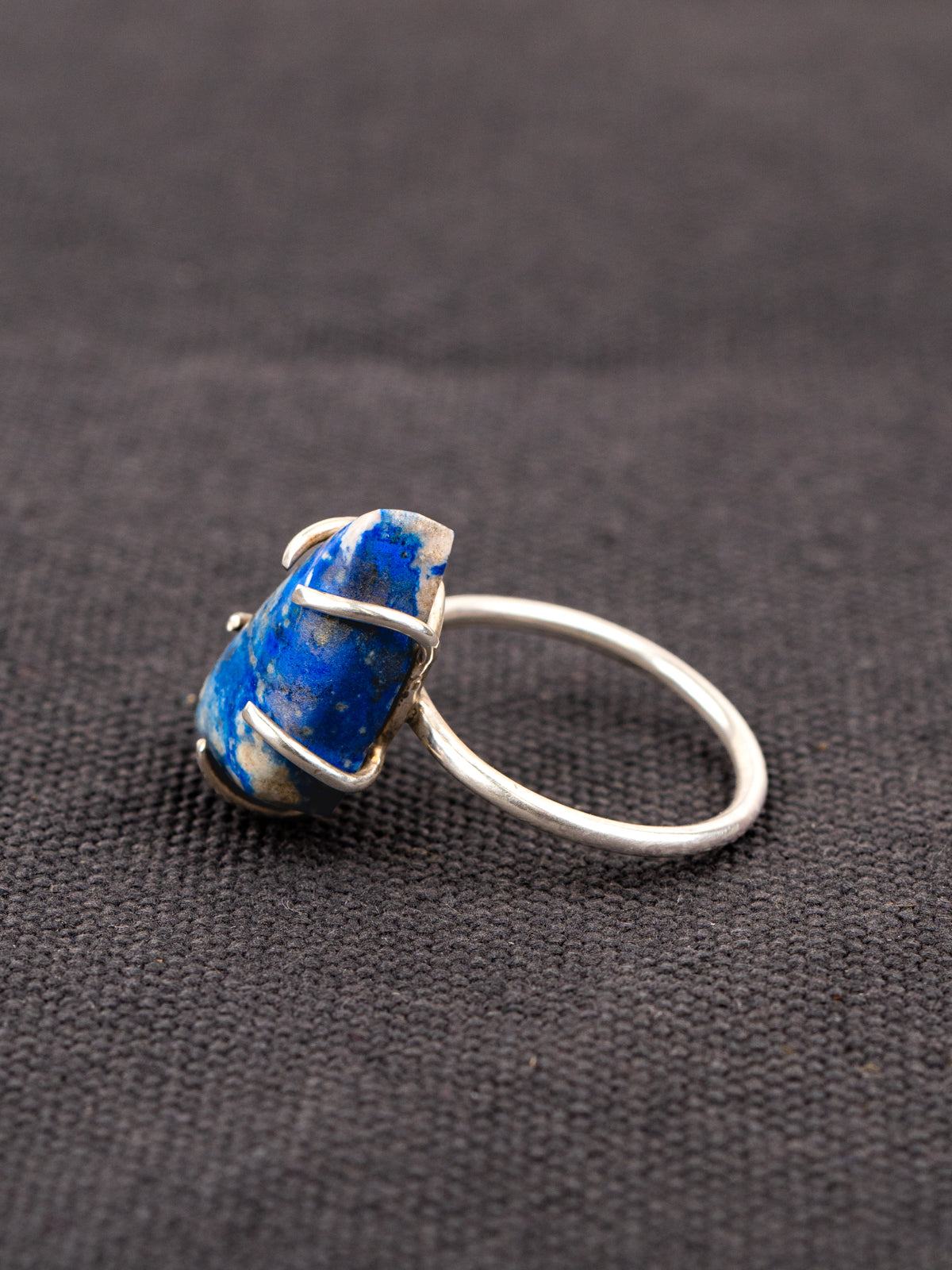 Side profile of a silver ring with a deep blue lapis stone held securely by a prong setting, displayed on a dark fabric background.
