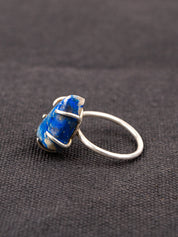 Side profile of a silver ring with a deep blue lapis stone held securely by a prong setting, displayed on a dark fabric background.
