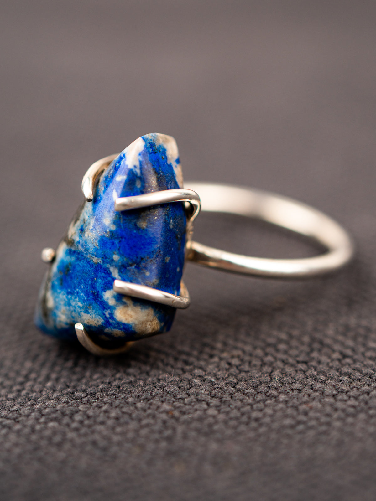 Up-close of the silver ring with a deep blue lapis stone held securely by a prong setting, displayed on a dark fabric background.