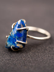 Up-close of the silver ring with a deep blue lapis stone held securely by a prong setting, displayed on a dark fabric background.
