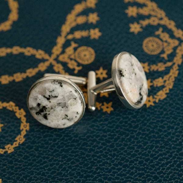Round sterling silver cufflinks set with a textured granite rock on a blue book with gold patterns.