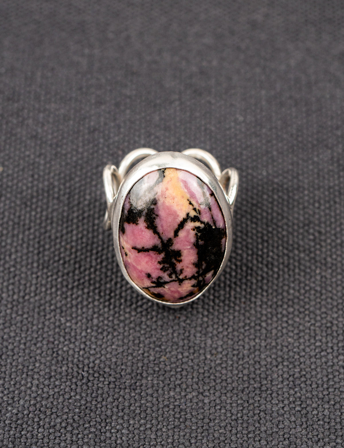Silver ring with a loop band design featuring a pink and black Rhodonite stone, displayed on a dark fabric background.