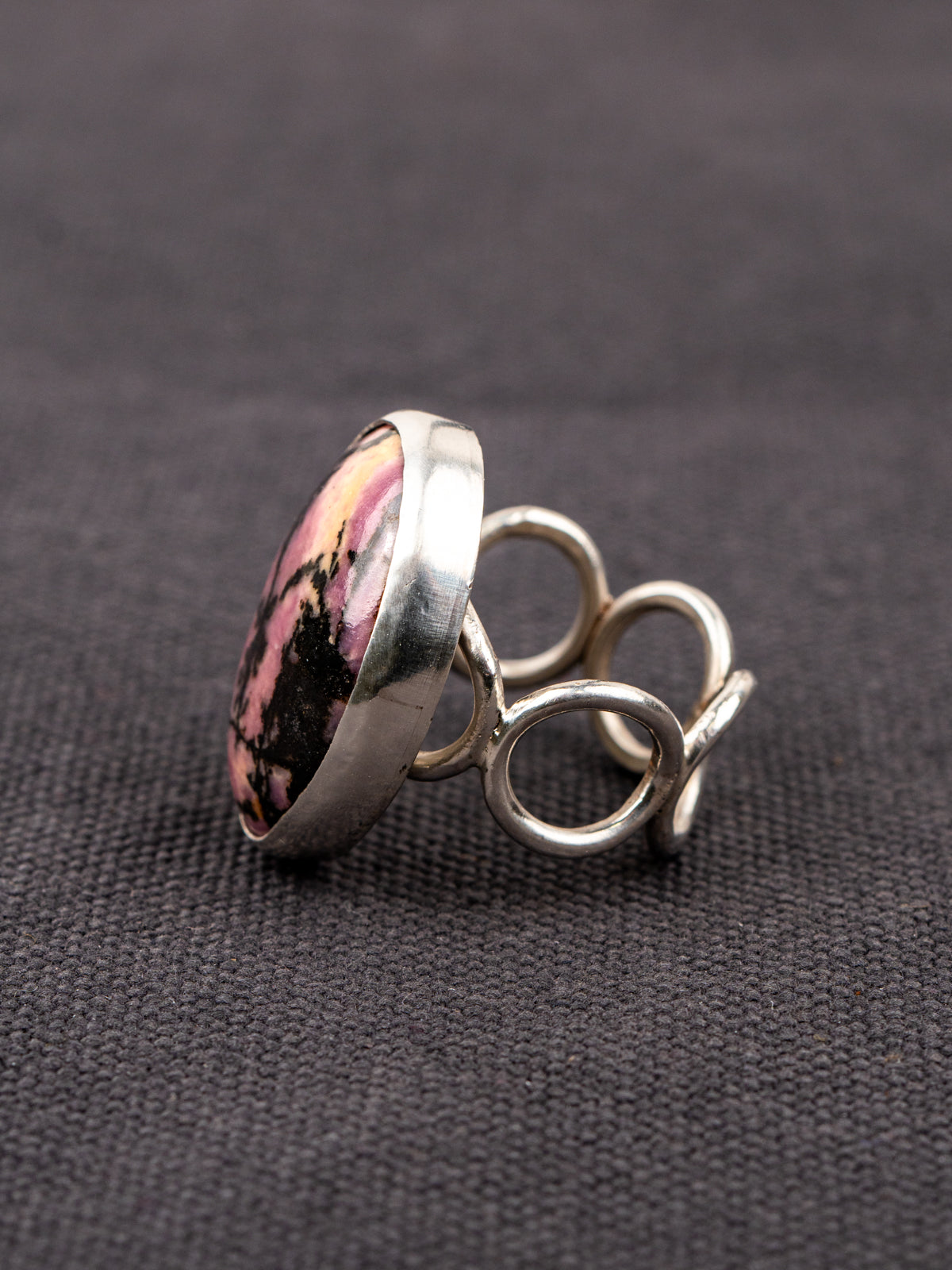 Side profile of a silver ring with a loop band design featuring a pink and black Rhodonite stone, displayed on a dark fabric background.