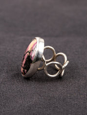 Side profile of a silver ring with a loop band design featuring a pink and black Rhodonite stone, displayed on a dark fabric background.