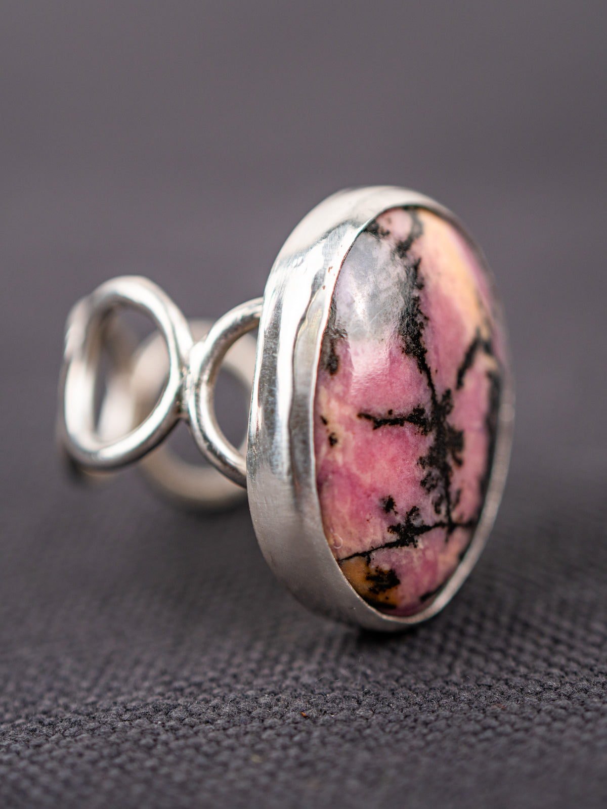 Close-up of a silver ring with a loop band design featuring a pink and black Rhodonite stone, displayed on a dark fabric background.