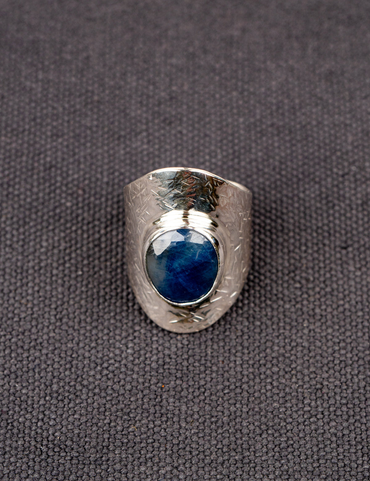 Silver statement ring with a wide textured band and a round deep-blue faceted Sapphire, set in a smooth bezel, displayed on a dark fabric background.