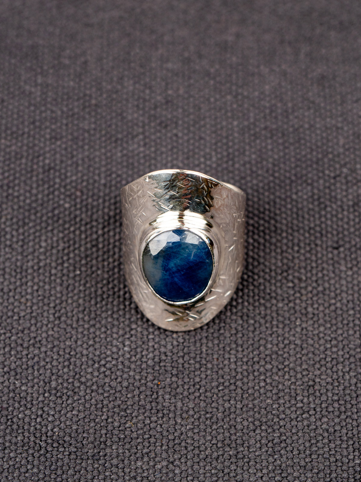 Silver statement ring with a wide textured band and a round deep-blue faceted Sapphire, set in a smooth bezel, displayed on a dark fabric background.