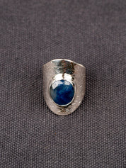 Silver statement ring with a wide textured band and a round deep-blue faceted Sapphire, set in a smooth bezel, displayed on a dark fabric background.