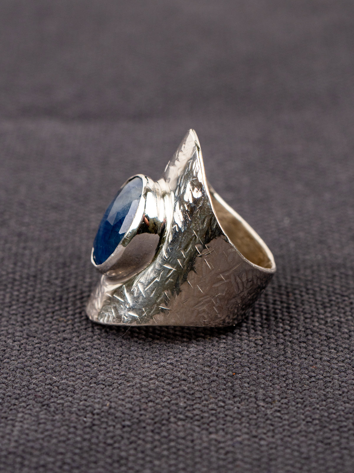 Silver statement ring with a wide textured band and a round deep-blue faceted Sapphire, set in a smooth bezel, displayed on a dark fabric background.