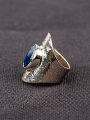Silver statement ring with a wide textured band and a round deep-blue faceted Sapphire, set in a smooth bezel, displayed on a dark fabric background.