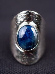 Up-close Silver statement ring with a wide textured band and a round deep-blue faceted Sapphire, set in a smooth bezel, displayed on a dark fabric background.
