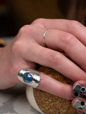 Silver statement ring with a wide textured band and a round deep-blue faceted Sapphire, set in a smooth bezel, displayed on a person's finger