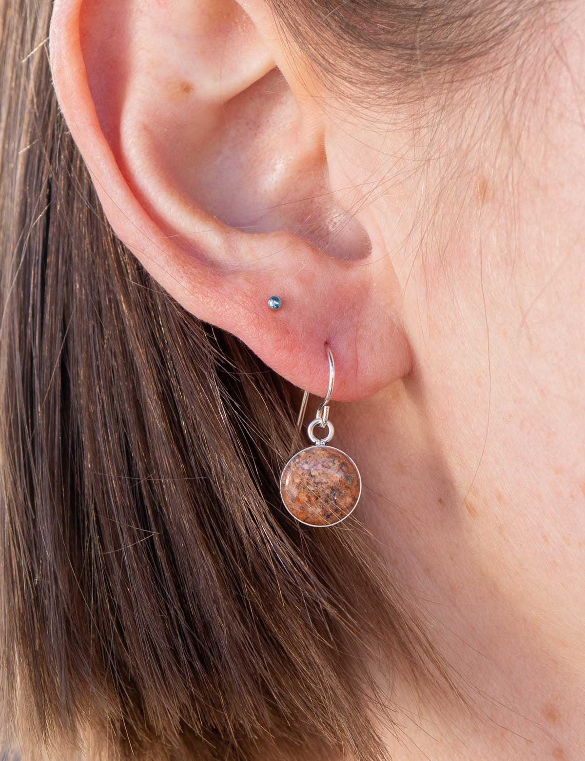 Close-up of an ear wearing a round dangle earring set with a chunk of natural red granite