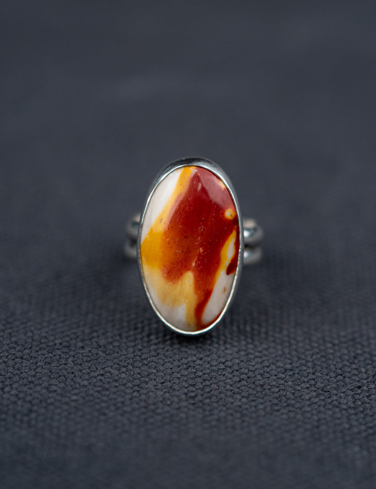 Mookaite Jasper ring featuring a smooth oval stone with warm red, yellow, and cream tones, set in a metal bezel with a shank band, displayed on dark fabric.