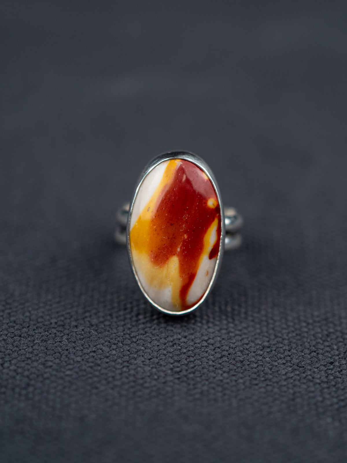 Mookaite Jasper ring featuring a smooth oval stone with warm red, yellow, and cream tones, set in a metal bezel with a shank band, displayed on dark fabric.