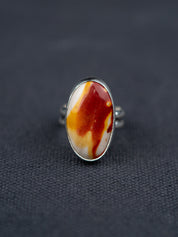 Mookaite Jasper ring featuring a smooth oval stone with warm red, yellow, and cream tones, set in a metal bezel with a shank band, displayed on dark fabric.