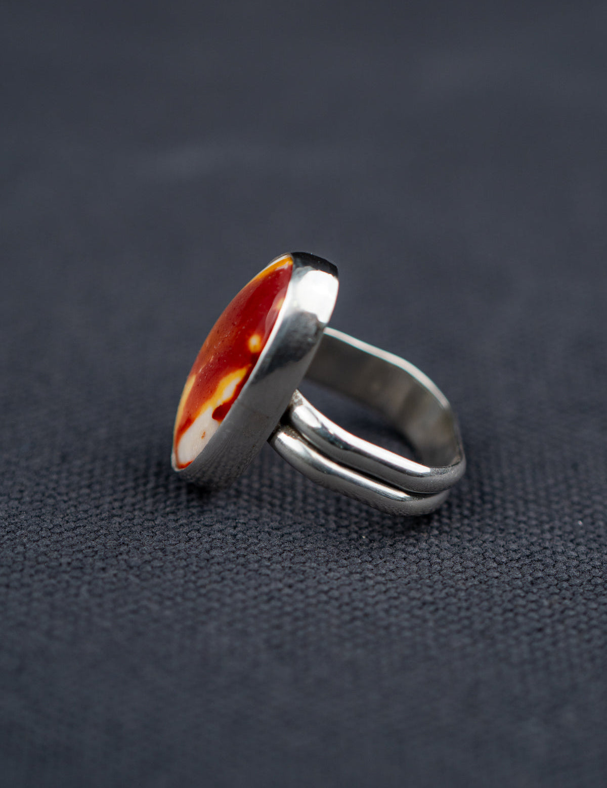 Side profile of a Mookaite Jasper ring featuring a smooth oval stone with warm red, yellow, and cream tones, set in a metal bezel with a shank band, displayed on dark fabric.