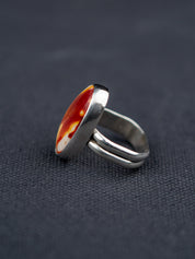 Side profile of a Mookaite Jasper ring featuring a smooth oval stone with warm red, yellow, and cream tones, set in a metal bezel with a shank band, displayed on dark fabric.