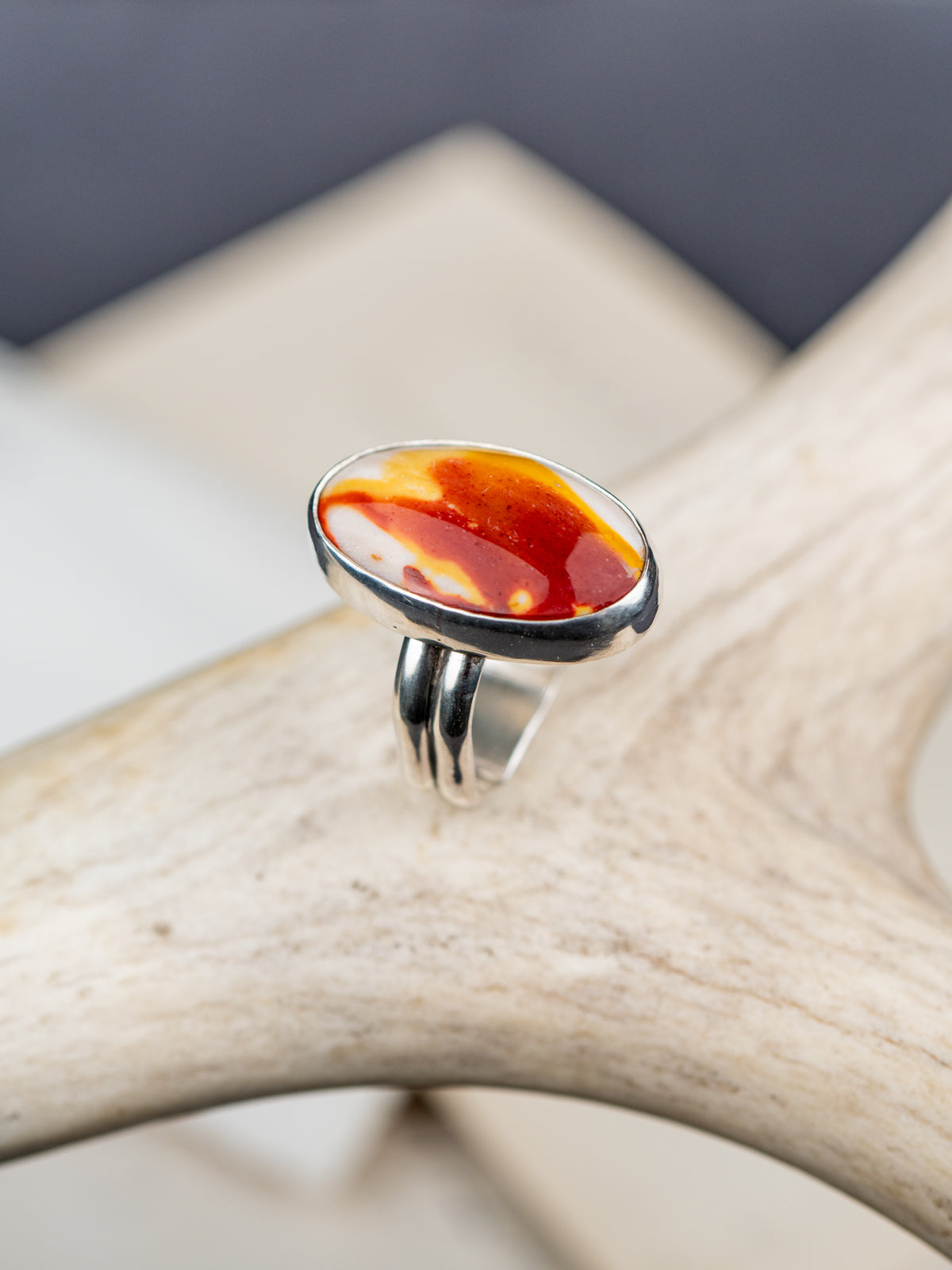 Mookaite Jasper ring featuring a smooth oval stone with warm red, yellow, and cream tones, set in a metal bezel with a shank band, displayed on a cream colored antler, with a dark fabric background.
