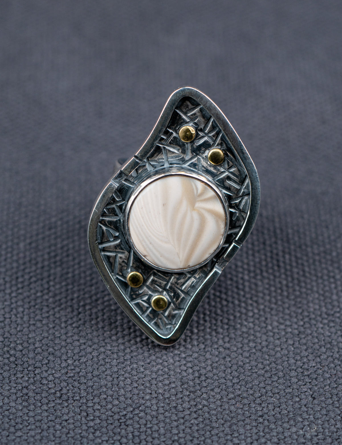 Silver ring with a textured S-curved-shaped metal frame, small brass studs, and a round cream-colored Flint stone, displayed on a dark gray fabric background.
