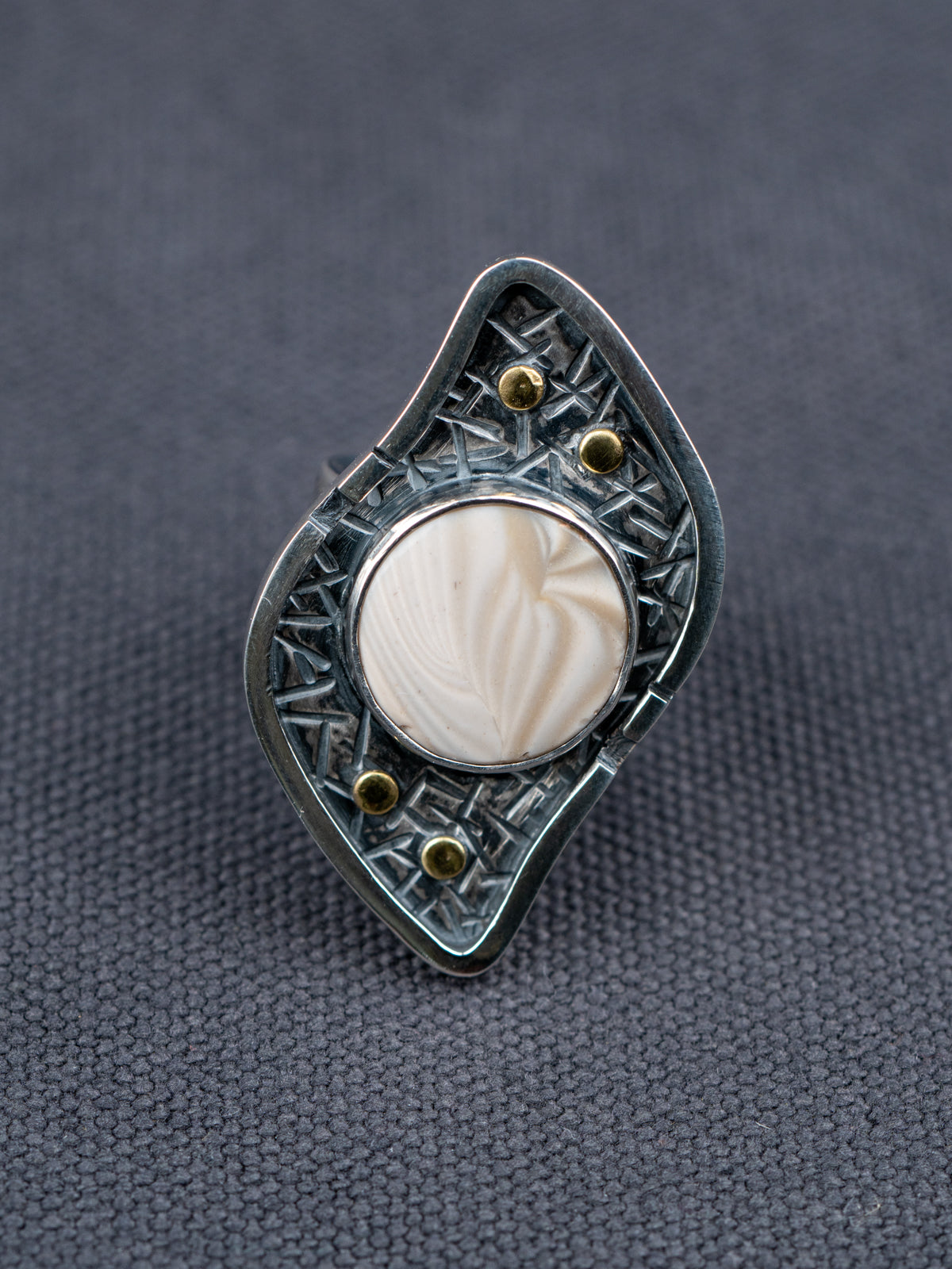 Silver ring with a textured S-curved-shaped metal frame, small brass studs, and a round cream-colored Flint stone, displayed on a dark gray fabric background.