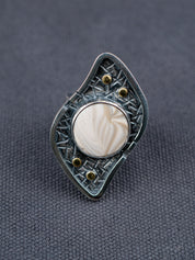 Silver ring with a textured S-curved-shaped metal frame, small brass studs, and a round cream-colored Flint stone, displayed on a dark gray fabric background.