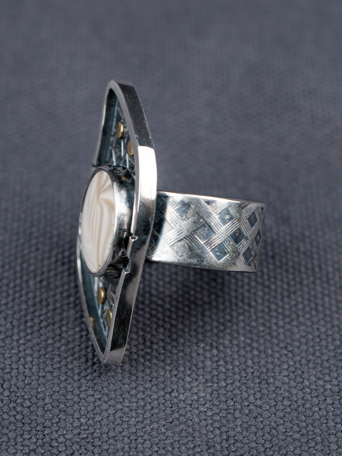 Side profile of a silver ring with a textured S-curved-shaped metal frame, small brass studs, and a round cream-colored Flint stone, displayed on a dark gray fabric background.