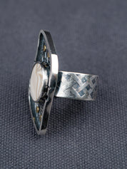 Side profile of a silver ring with a textured S-curved-shaped metal frame, small brass studs, and a round cream-colored Flint stone, displayed on a dark gray fabric background.