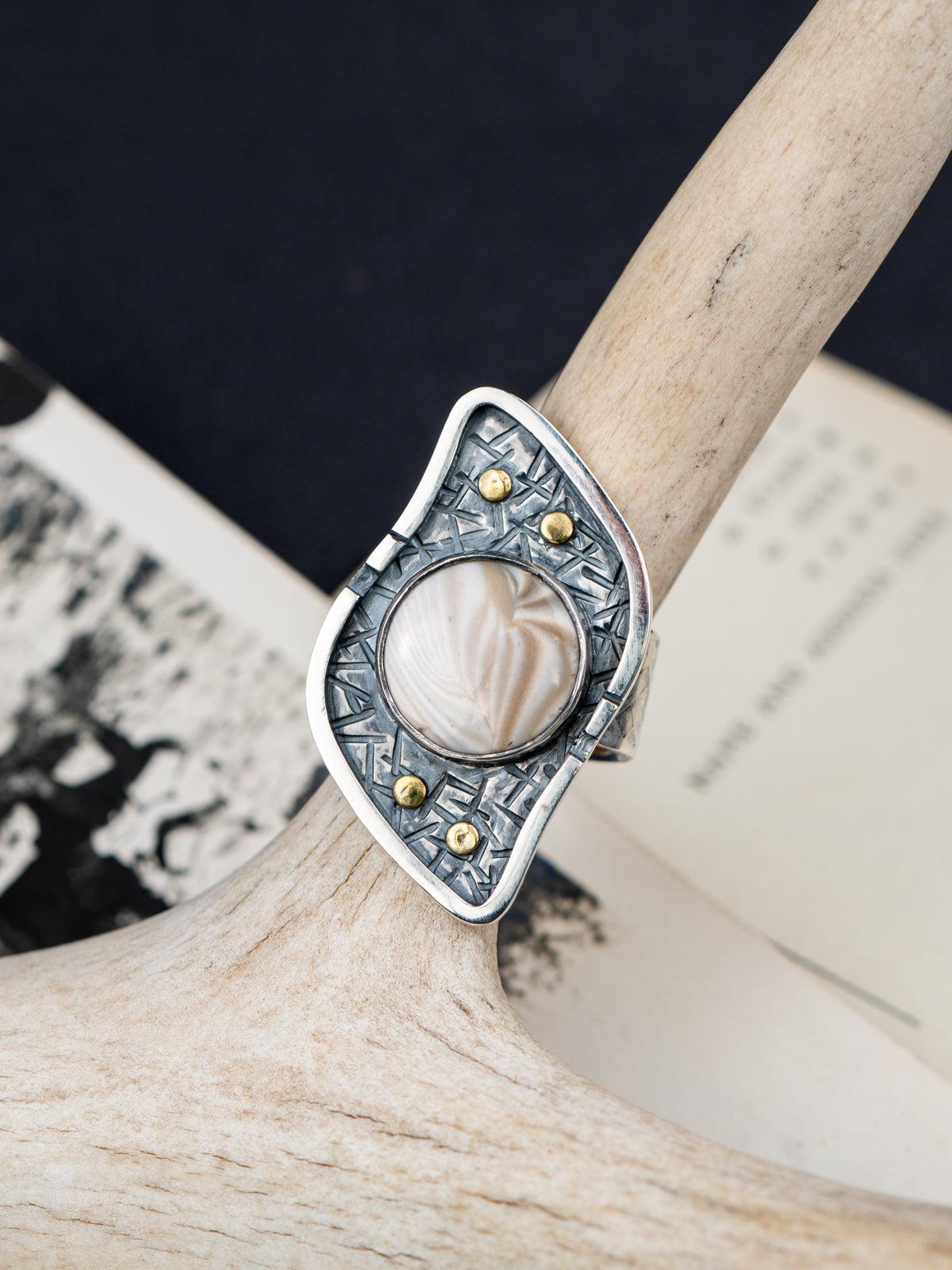 Silver ring with a textured S-curved-shaped metal frame, small brass studs, and a round cream-colored Flint stone, displayed on antlers with postcards in the background.