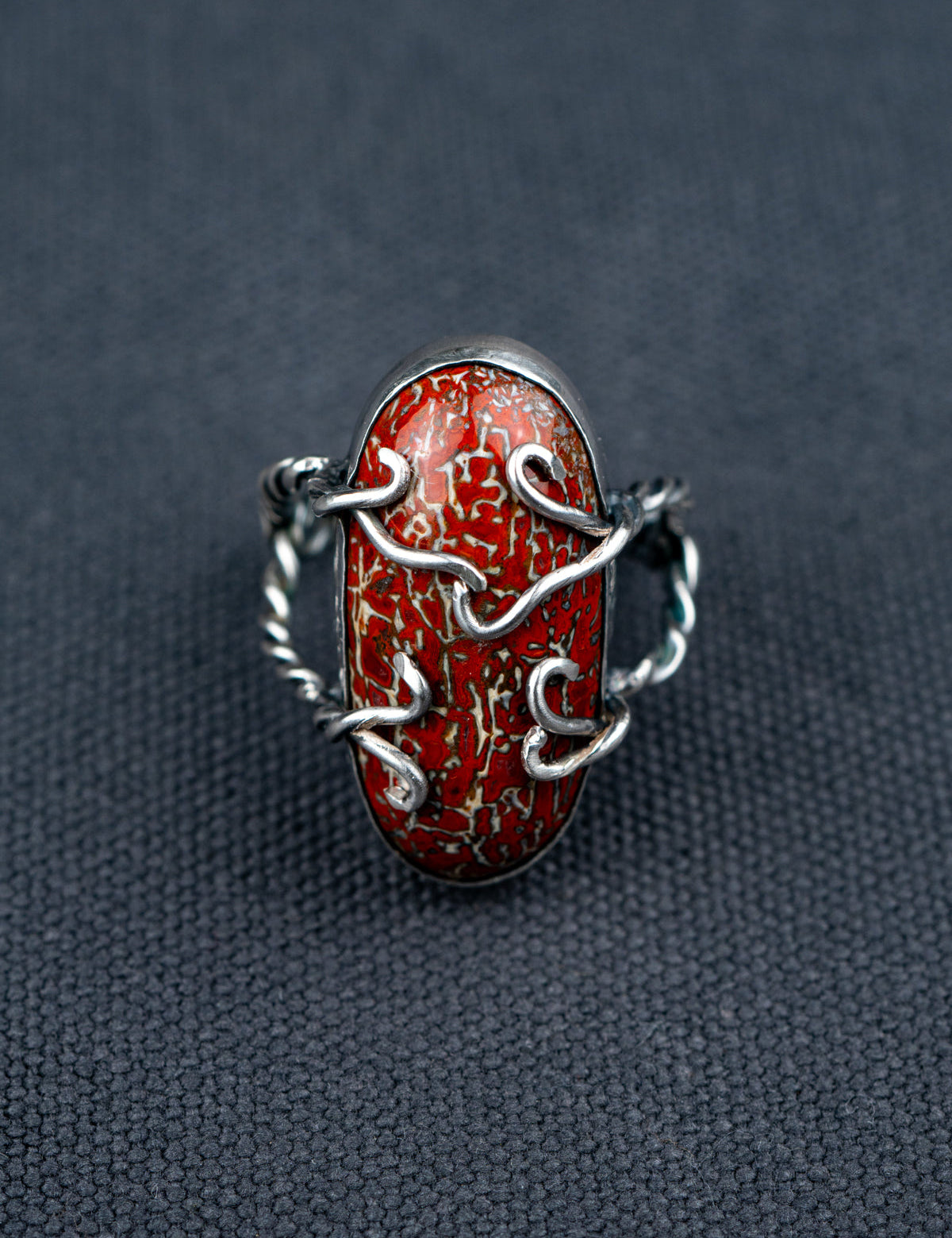 Dinosaur bone stone ring featuring a patterned fossilized gemstone set in a metal bezel with a twisted band, displayed on dark textured fabric.