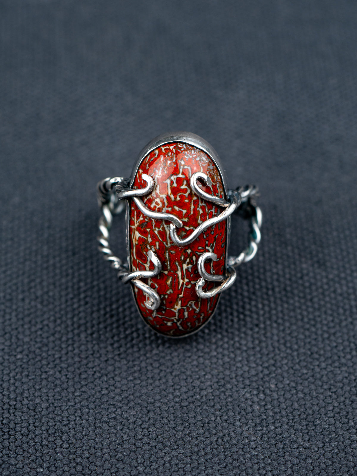 Dinosaur bone stone ring featuring a patterned fossilized gemstone set in a metal bezel with a twisted band, displayed on dark textured fabric.