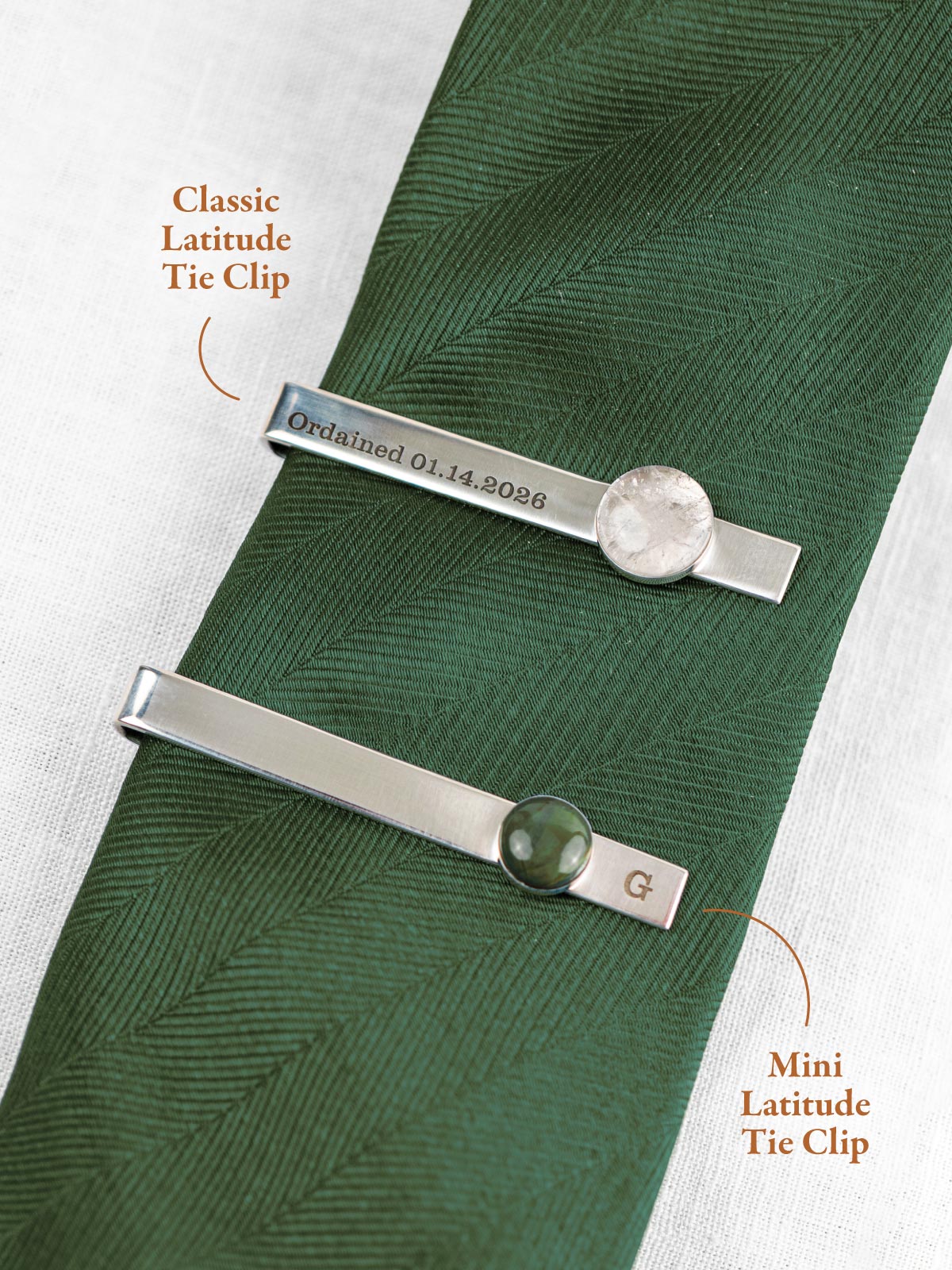 Two tie clips on a green tie with text indicating the size of the round stone set on the clip, the larger 'Classic Latitude Tie Clip' and the smaller 'Mini Latitude Tie Clip'